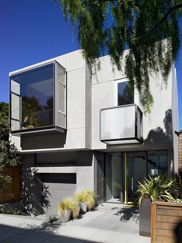 Exterior view of a compact contemporary multi-storey residence with a white rendered and concrete facade, projecting glass-enclosed upper-level window, projecting box window, dark garage door, and glazed entry. The building sits along a paved frontage with timber boundary fencing and large planted containers, viewed in bright sunlight beneath a clear blue sky.