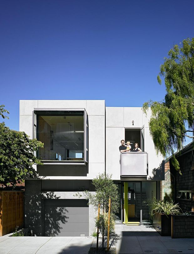 Front exterior view of a compact multi-storey urban residence with a minimalist cubic form. The facade combines smooth white rendered panels with dark lower-level cladding, a recessed garage, large glazed openings, and a projecting upper-floor balcony. A bright green entry door provides a vivid accent, while mature planting and neighboring residences frame the narrow landscaped approach.