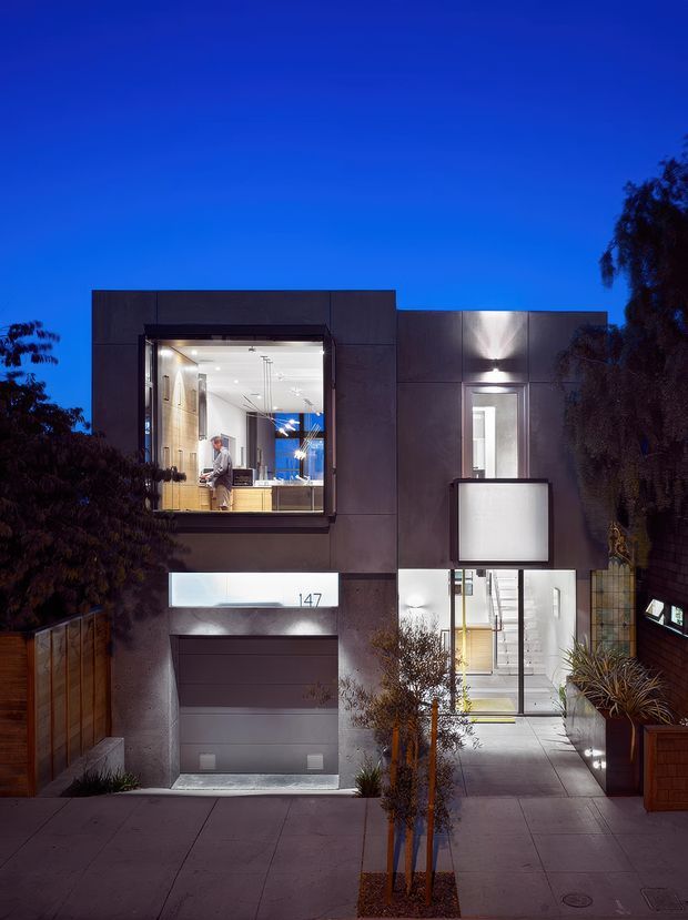 Twilight exterior photograph of a narrow contemporary multi-storey urban residence. The dark, rectilinear facade has large illuminated windows, a recessed entry, a frosted projecting feature panel, integrated exterior lighting, a garage, concrete paving, and timber fencing. Interior spaces are visible through the glazing, with a person standing near a kitchen or living area.