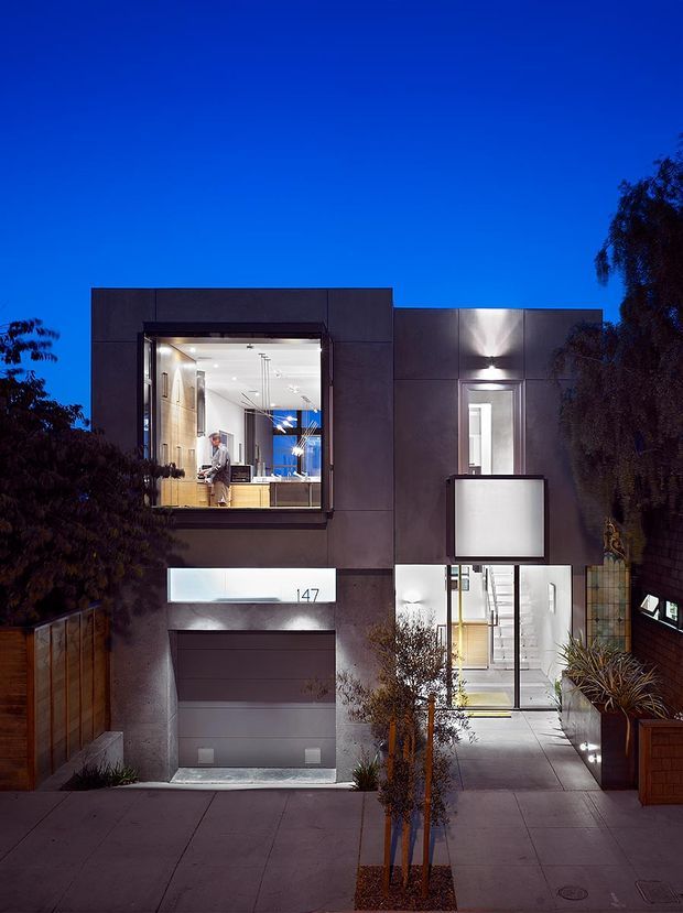 Twilight exterior photograph of a contemporary multi-storey urban residence. The dark, rectilinear facade features large illuminated windows, a recessed entry with a projecting box-like canopy, an integrated garage, and a paved front approach with low planting and timber boundary fencing. Interior activity and cabinetry are visible through the glazing.