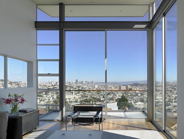Bright, high-level interior with floor-to-ceiling corner glazing framing a broad urban skyline and distant water. The room includes a small seating area, a low coffee table, a side table with flowers, and exposed dark structural window framing. Strong daylight, expansive views, and a compact furnished lounge create an airy contemporary atmosphere.