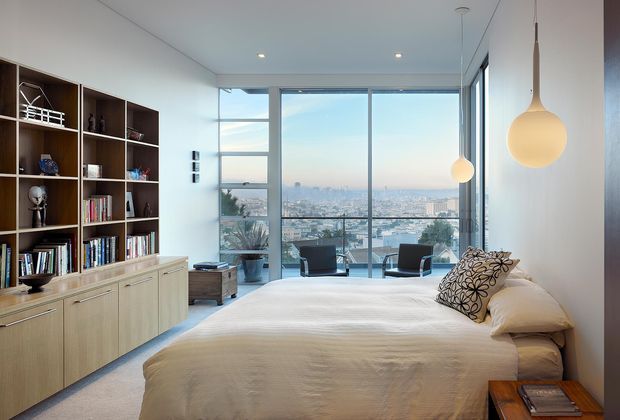 A bright contemporary bedroom with a large bed in the foreground, built-in pale timber shelving and cabinetry along the left wall, and pendant lights beside the bed. Floor-to-ceiling glazing opens to a balcony or terrace with expansive city and water views. The room has a clean, uncluttered design and abundant natural light.