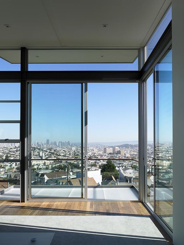 Bright, minimally furnished interior corner with expansive floor-to-ceiling glazing and dark-framed sliding doors, opening toward a balcony or terrace. The elevated view overlooks a dense urban neighbourhood and distant city skyline. The room has a pale smooth ceiling, light neutral walls, and a mixed light grey and warm-toned floor finish, with strong sunlight and long shadows.