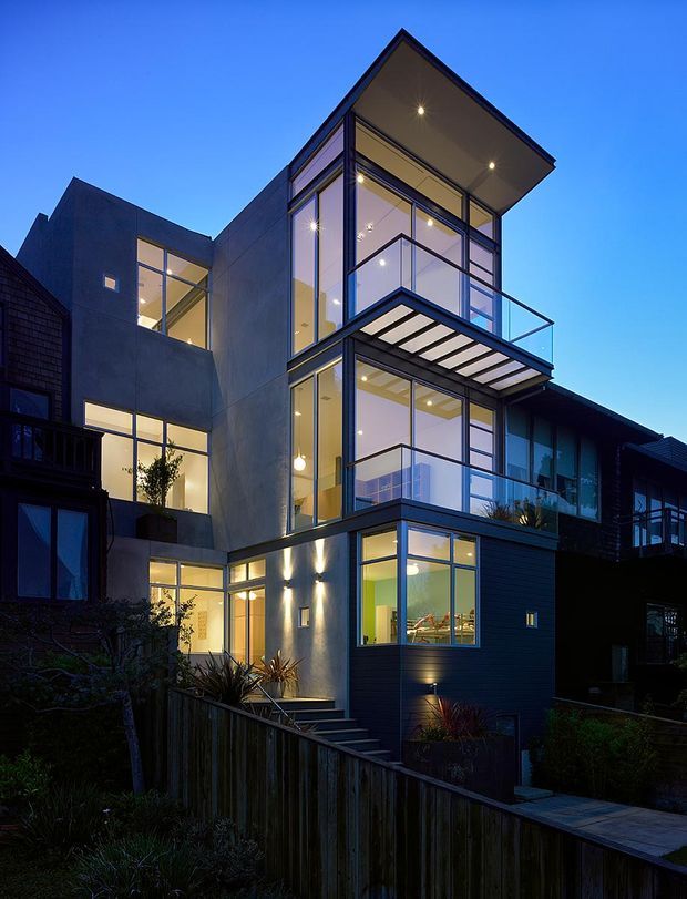Dusk exterior view of a contemporary multi-storey urban residence, featuring a tall glazed central volume, projecting balconies with clear glass balustrades, dark horizontal cladding, smooth concrete-like wall surfaces, and extensive illuminated windows. A sloping landscaped site and timber boundary fencing are visible in the foreground.