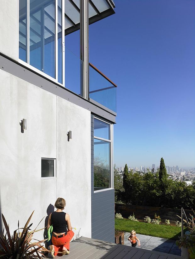 A modern multi-level residential exterior with crisp white rendered walls, dark horizontal cladding, expansive floor-to-ceiling glazing, and glass balustrades. The building opens onto a landscaped garden and deck, with a distant urban skyline visible beyond the property. Strong geometric lines, projecting roof canopies, and abundant glazing create a contemporary architectural character.