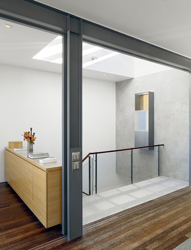 Bright contemporary interior showing a stair landing and hallway viewed through a large dark-framed glazed partition. The space features a white ceiling with skylights, a textured pale grey feature wall, a recessed vertical display niche, glass balustrades with dark timber handrails, white stair treads, and a light timber built-in sideboard adjoining a dark timber floor and striped carpeted landing.