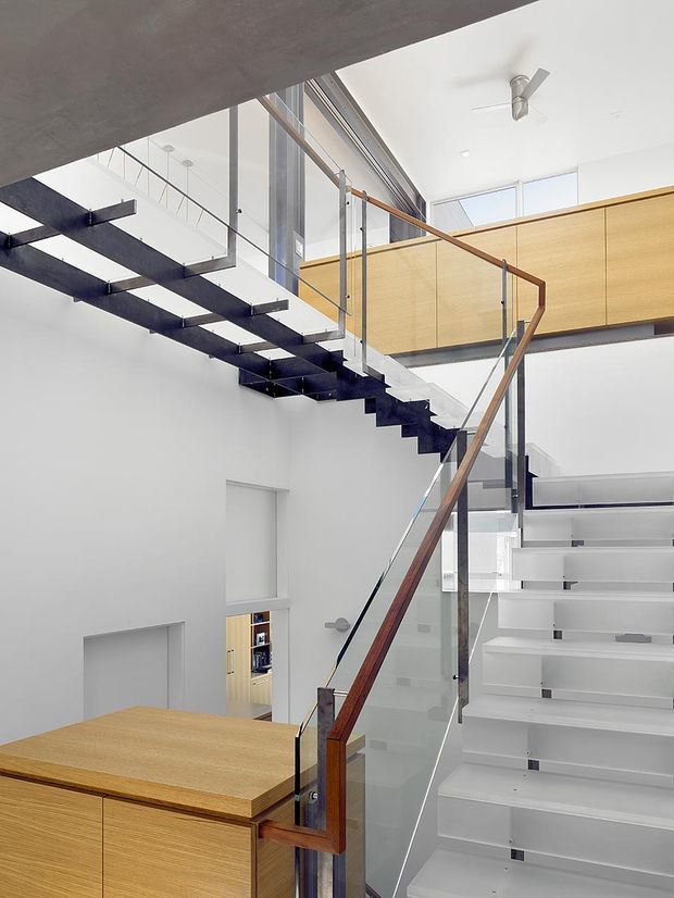 Bright modern multi-level stairwell with sculptural open-riser stairs, clear glass balustrades, dark steel stringers, and warm timber handrails and cabinetry. A compact kitchen or joinery area is visible below, while an upper-level timber-lined mezzanine or bridge overlooks the void. The space has white walls and ceilings, strong vertical glazing, and a clean contemporary architectural character.