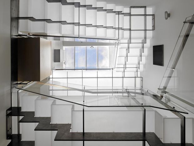 Interior view of a contemporary multi-level stairway and landing with a dramatic double-height void. Multiple flights of open-riser stairs, dark timber or stone treads, clear glass balustrades with slim dark metal supports, white walls, and extensive glazing create a bright, highly geometric space. Fixed dark cabinetry is visible at the left, with wall-mounted screens or panels on the right.