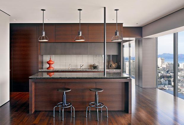 Contemporary high-rise apartment kitchen with a large dark timber island, integrated dark cabinetry, stone benchtops, tiled splashback, and three pendant lights. Two bar stools sit at the island, while floor-to-ceiling glazing provides expansive city and mountain views. The room has a refined, warm, high-contrast palette and polished modern detailing.