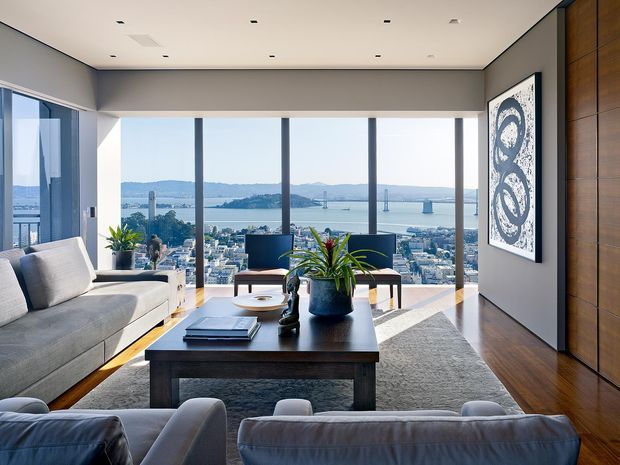 A refined contemporary living room with floor-to-ceiling glazing overlooking a dense waterfront cityscape and suspension bridge. The room features a large grey sectional sofa, dark timber coffee table, grey area rug, low lounge chairs, indoor plants, recessed ceiling lighting, and a full-height dark timber feature wall with framed artwork.