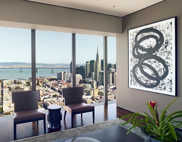 A refined high-rise interior with floor-to-ceiling glazing overlooking a dense city skyline and bay. Two upholstered lounge chairs and a small side table sit on dark timber flooring, while a stone-topped counter or island occupies the foreground. Neutral grey walls, a large monochrome abstract artwork, and indoor greenery create a contemporary, polished atmosphere.
