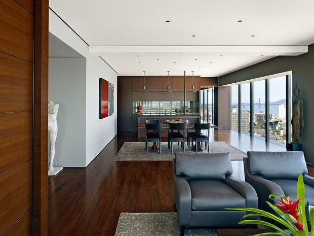 A sophisticated open-plan apartment interior combining a living room and dining area, with dark timber flooring, warm timber wall panelling and cabinetry, a glass-fronted kitchen, and floor-to-ceiling glazing overlooking a dense city and waterfront skyline. A grey sectional sofa and armchairs occupy the foreground, while a dining table sits centrally beneath a broad recessed ceiling detail. The palette is dark and warm with cool grey furnishings, and the overall aesthetic is refined contemporary urban design.