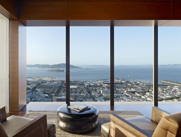 A refined modern interior lounge or viewing room framed by extensive floor-to-ceiling glazing, overlooking a coastal harbour, waterfront city and distant hills. Warm timber ceiling and wall lining contrast with dark vertical window mullions, pale upholstered lounge chairs, and a low glossy black round coffee table on a textured neutral rug. The composition is calm, airy and strongly focused on the expansive water view.
