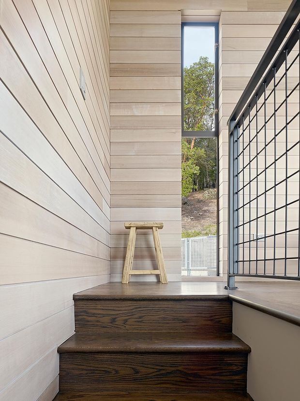 A narrow, modern interior stairway with dark timber stair treads and risers, pale horizontal timber wall panelling, and a black metal-framed balustrade. A tall vertical window brings in natural light and overlooks trees and a sloping outdoor site. A small timber stool sits on the landing.