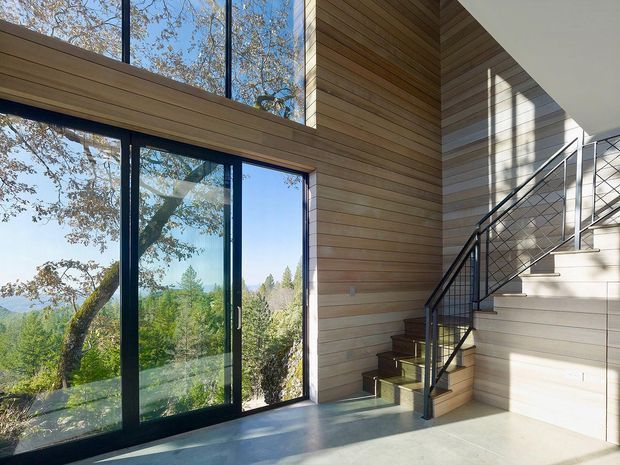Bright contemporary interior stair hall with a broad black-framed glazed opening overlooking trees and a distant landscape. The space features extensive horizontal timber wall and ceiling lining, a polished light-grey concrete floor, and a staircase with dark timber treads and a slender black metal balustrade. Strong natural light creates a calm, warm, nature-connected atmosphere.