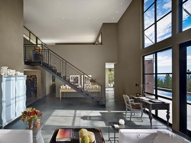A spacious double-height open-plan living area with a dramatic wall of floor-to-ceiling glazing overlooking a landscape. A steel-framed open-riser staircase with wire balustrade crosses the room, while a traditional dark wood desk sits beside the windows. The room combines polished concrete flooring, warm neutral walls, contemporary recessed lighting, and a mix of classic and modern furnishings.