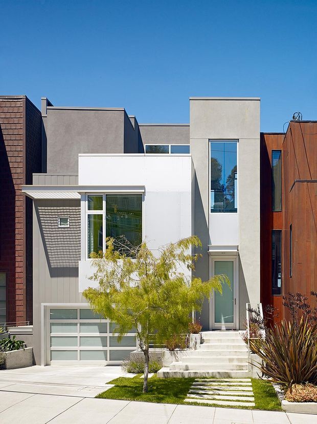 Front exterior view of a contemporary multi-storey urban townhouse. The facade combines smooth grey and white rendered volumes with dark vertical cladding, warm rust-toned metal cladding, tall narrow windows, a projecting entry canopy, and an integrated garage. A paved driveway and stepped concrete path cross a small landscaped front garden with lawn and sculptural planting under a clear blue sky.