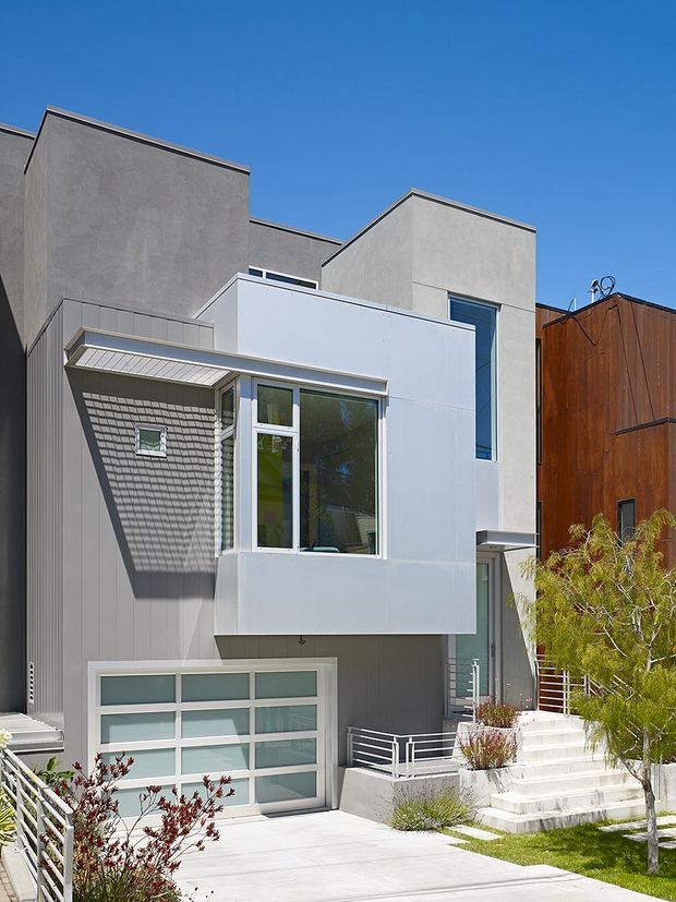 Exterior view of a contemporary multi-level urban residence with intersecting box-like volumes, flat rooflines, smooth grey stucco walls, white metal-clad projecting elements, vertical glazing, and a frosted-glass sectional garage door. A covered entry canopy, white metal railings, concrete steps, driveway, and a small landscaped front garden are visible. Strong sunlight creates pronounced shadows against a clear blue sky.