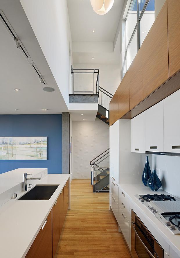 Bright contemporary kitchen with white benchtops, timber cabinetry, and integrated white joinery, opening into a tall double-height circulation space with a metal-framed staircase and upper-level landing. Large vertical windows bring in abundant daylight, while a blue feature wall and blue glass vessels provide colour contrast.