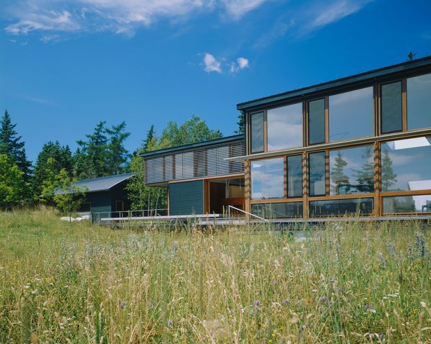 Exterior view of a contemporary two-storey house set in a natural meadow, with extensive floor-to-ceiling glazing, dark horizontal cladding, timber-framed windows, and a projecting upper-level volume. A separate dark-roofed outbuilding is visible to the left, surrounded by mature trees and wild grasses.