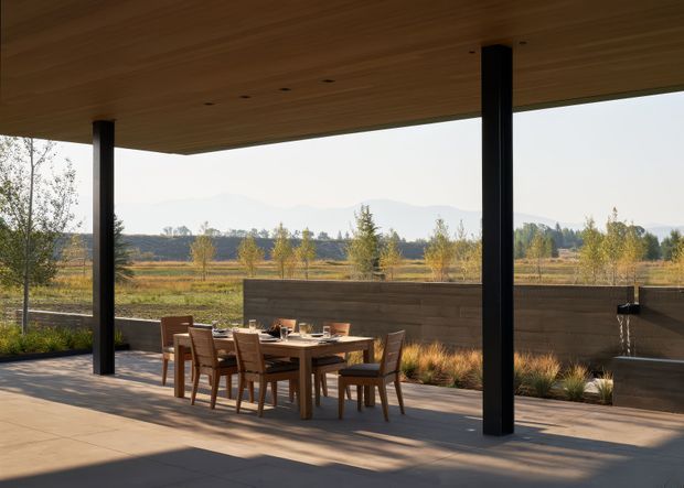 A covered outdoor dining terrace with a timber-lined flat roof supported by slender black columns. A timber dining table and chairs sit on a broad concrete-paved floor, beside a low dark timber-clad wall and a minimalist water feature. The open view looks across landscaped grounds toward distant hills, creating a calm, contemporary indoor-outdoor setting.