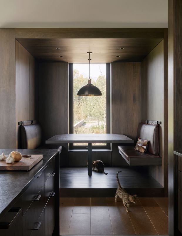 A compact, dark-toned dining nook integrated beside a kitchen, with floor-to-ceiling dark timber wall and ceiling panelling, built-in banquette seating, a central dining table, and a black dome pendant light. A large vertical window brings in natural light and overlooks greenery. Dark kitchen cabinetry and a stone benchtop are visible in the foreground, with cats on the floor and seating.