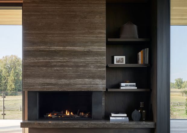 A refined contemporary living room feature wall centred on a linear fireplace with flames, framed by dark built-in shelving and cabinetry. The fireplace surround and lower hearth use layered dark brown stone or timber-toned material, while open shelves display books and understated objects. Large windows on both sides provide views to a rural landscape, and warm roller blinds soften the daylight.