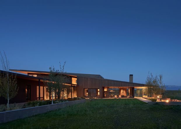 A low, sprawling contemporary residence photographed at dusk, with warm interior lighting glowing through extensive glazing. The house has a largely flat or very low-pitched roof, dark timber-toned horizontal cladding, projecting roof eaves, and a long linear composition opening onto a broad lawn and paved outdoor area. Young trees and planted garden beds frame the building, while distant terrain is visible beyond the site.