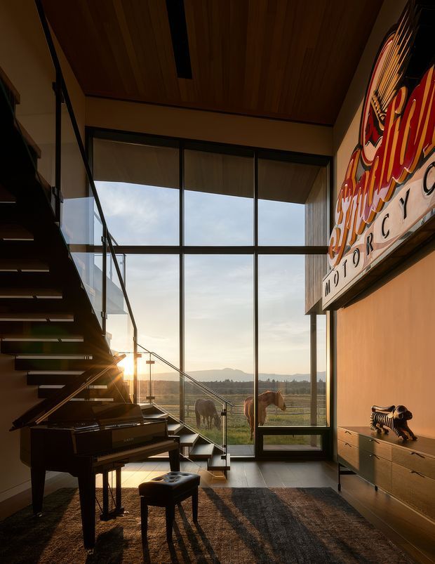 A dramatic double-height living space with a grand piano, floating open-riser staircase, expansive black-framed floor-to-ceiling glazing, timber ceiling, and built-in timber cabinetry. Warm late-afternoon light enters through the glazing, with horses visible outside in the rural landscape. A large colourful graphic artwork occupies the upper wall, creating a bold focal point.