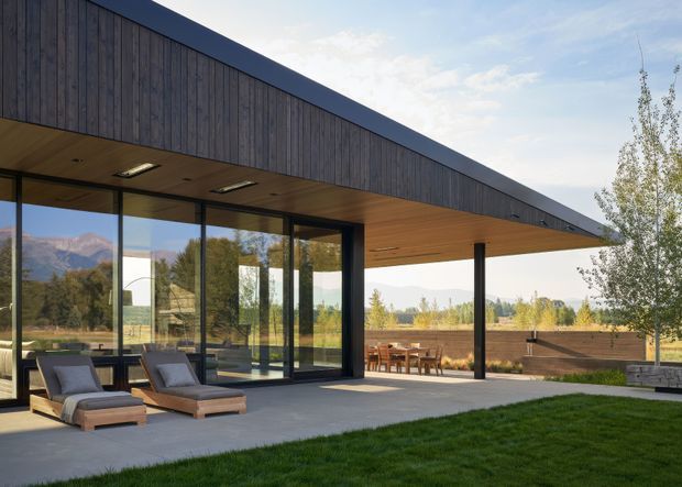 A modern single-storey pavilion-style home with a long dark timber-clad roof volume, deep timber-lined eaves, extensive floor-to-ceiling glazing, and slender dark structural columns. The covered terrace contains outdoor dining furniture and lounge chairs, opening onto a manicured lawn with mountain and rural landscape views beyond.