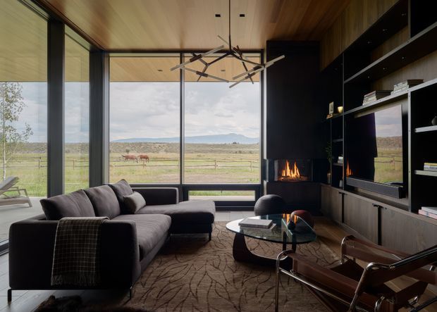 A sophisticated living room with floor-to-ceiling glazing overlooking an open rural landscape. The room features a large charcoal sectional sofa, sculptural glass-and-metal coffee table, leather lounge chair, dark built-in media cabinetry, and a linear fireplace. Warm timber covers the ceiling and upper wall surfaces, while a statement branching pendant hangs centrally.