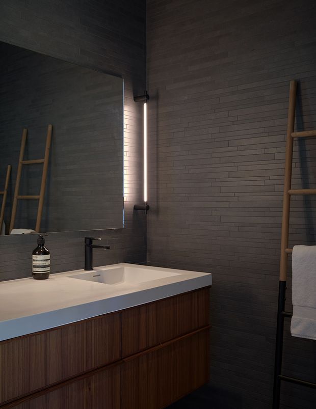 A moody contemporary bathroom vanity with a white integrated basin and countertop, dark timber vertical-grain cabinetry, charcoal grey brick-pattern wall tiles, a large mirror with linear wall lighting, and a timber ladder-style towel rail.