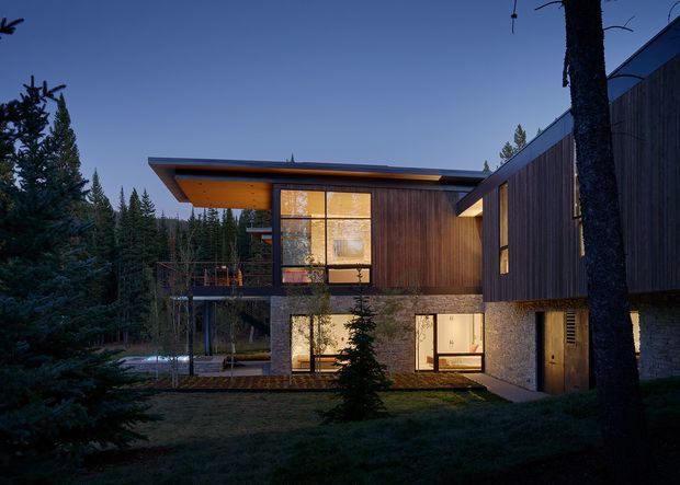 Exterior architectural photograph of a contemporary multi-level house set within a dense forest at dusk. The home features strong horizontal roof planes, extensive floor-to-ceiling glazing, warm timber soffits and vertical timber cladding, dark structural framing, and a textured masonry wall at the lower level. Warm interior lighting contrasts with the cool blue evening light and surrounding evergreen trees.