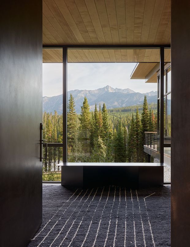 A dark, refined interior opening onto a dramatic mountain and forest landscape. Floor-to-ceiling glazing frames the view, with a deep black built-in bench or low cabinet in front of the window. The space features dark walls, a linear wood-plank ceiling, charcoal carpet with thin light stripes, and a projecting exterior balcony visible to the right.