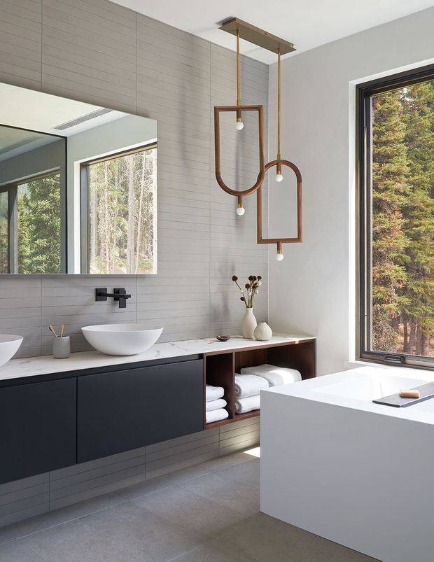 Bright contemporary bathroom featuring a floating dark vanity with open timber towel storage, a white vessel basin, pale horizontal wall tiles, large mirror, freestanding white bathtub, and sculptural suspended pendant lights. A large window provides views to dense woodland outside.