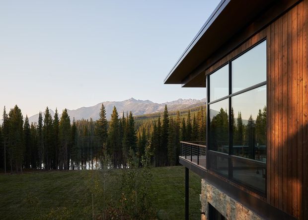 Exterior view of a contemporary mountain dwelling set among dense evergreen forest, with a projecting roof, dark timber-clad facade, expansive floor-to-ceiling glazing, a narrow balcony, and a stone-clad lower wall. Mountain ridges and a glimpse of water are visible beyond the trees.
