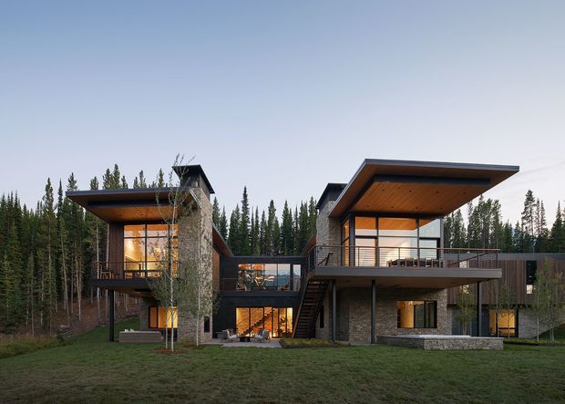 A contemporary multi-level house set within a dense forest, viewed from the front across a lawn. The building is composed of several connected volumes with broad cantilevered flat roof planes, extensive glazing, dark timber soffits, stone-clad walls, and elevated terraces. A central glazed link and exterior stair connect the wings, while warm interior lighting contrasts with the cool forest setting.