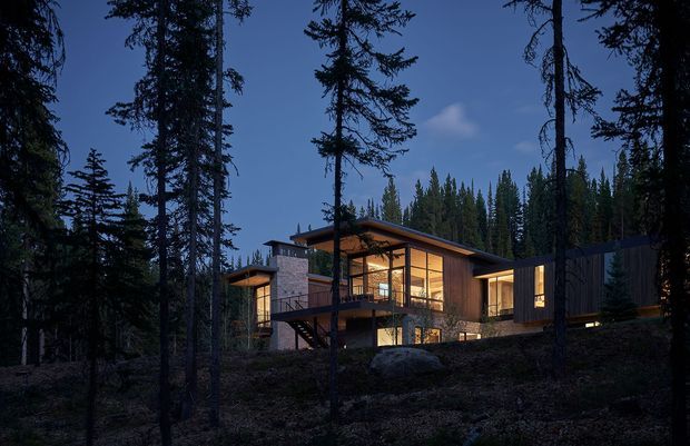A modern, single-level house set among a dense conifer forest at dusk. The dark-clad structure has broad floor-to-ceiling glazed sections glowing with warm interior light, a low-pitched roof with pronounced overhangs, a stone chimney or feature wall, and an elevated timber deck projecting over a sloped, rocky site.