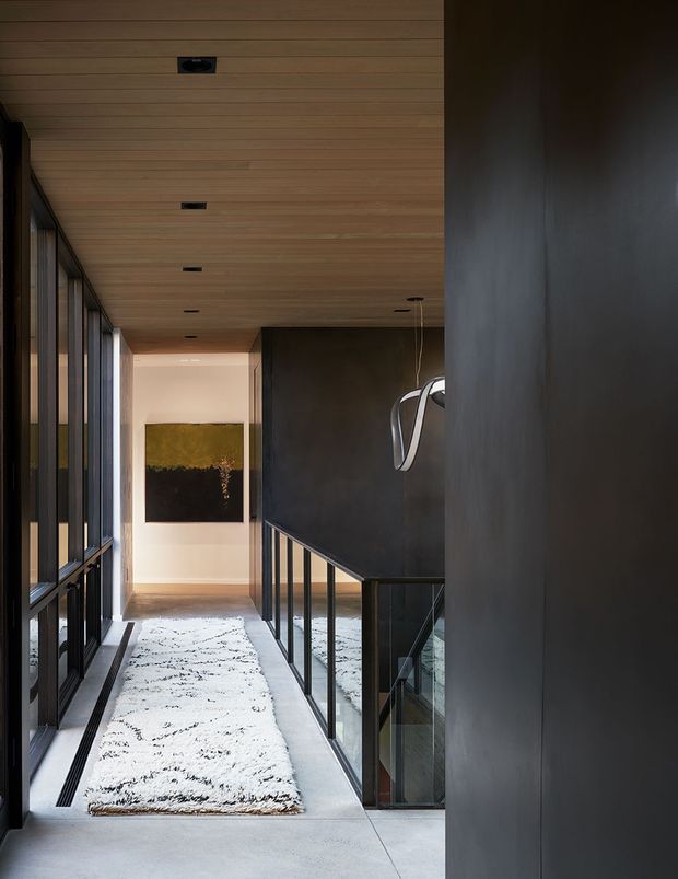 A sophisticated modern interior hallway or landing with a long white patterned rug over a smooth pale floor, dark-framed floor-to-ceiling glazing along the left side, and a black-framed glass balustrade overlooking a stairwell on the right. The ceiling is clad in horizontal light timber boards with recessed downlights, while dark charcoal walls and a sculptural pendant create a dramatic, refined atmosphere. Abstract artwork is visible at the far end.