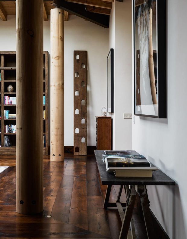 A narrow, warm-toned interior passage or living area with polished dark timber flooring, exposed timber ceiling beams, and prominent round natural timber structural columns. A dark built-in bookshelf filled with books occupies the left side, while a slim console table with books sits along the white wall on the right beneath framed artwork. A rustic vertical timber feature with arched cut-outs and a small timber cabinet add handcrafted character.