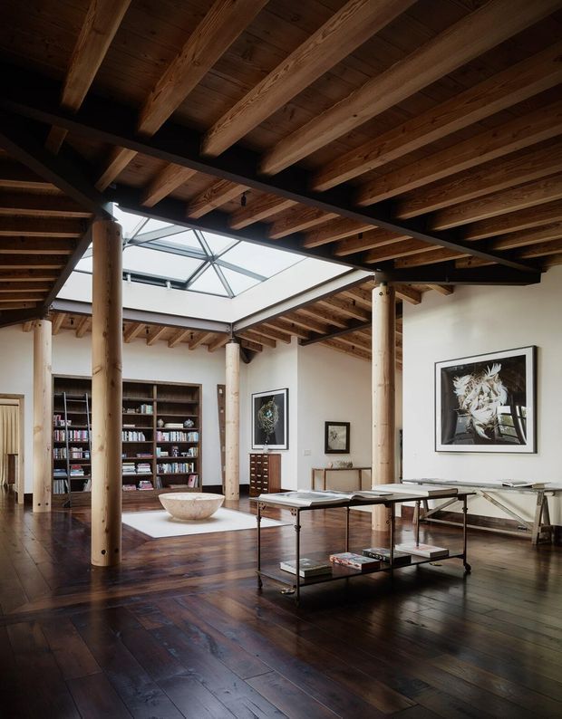 A spacious, moody open-plan interior with exposed timber roof beams and dark timber flooring. Thick round timber posts and black steel beams support the high ceiling, while a large central skylight brings in daylight. The room includes a built-in library, framed artwork, a low glass-and-metal coffee table, and a long glass-topped work or display table. The architecture has a warm timber-and-steel character with an industrial, loft-like feel.