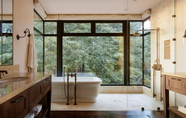 A spacious, refined bathroom with a freestanding white bathtub positioned in front of expansive dark-framed windows overlooking dense greenery. The room includes a double vanity with dark timber cabinetry and stone benchtops, a glass-enclosed shower with brass fixtures, pale tiled flooring and walls, and warm brass lighting and hardware.