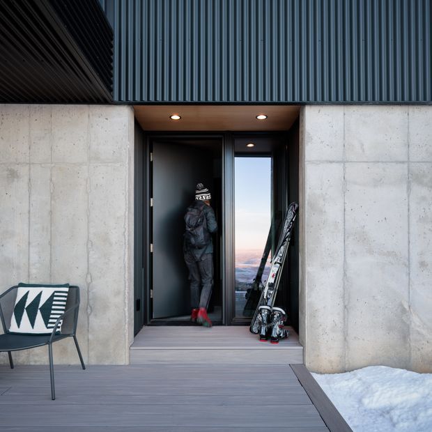 A modern mountain-home entry viewed from the exterior, with a dark metal-clad facade, raw concrete walls, recessed ceiling lights, a black entry door and narrow window, timber decking, ski equipment, and snow visible at the threshold. The doorway opens into a dark interior, with a distant mountainous view through the glazing.