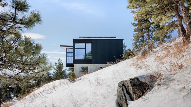 A modern, compact dark-clad house set into a steep, snow-covered, wooded hillside. The building has a flat roof, strongly rectilinear form, large glazed openings, and projecting window bays. Evergreen trees and exposed rock surround the elevated site.