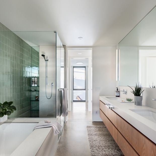 Bright contemporary bathroom with a freestanding bathtub in the foreground, a glass-enclosed walk-in shower lined with green square tiles, and a long light-wood vanity with a white countertop and integrated basins. Large wall mirrors, white walls and ceiling, pale flooring, recessed lighting, and a narrow windowed opening create an airy, clean space. A potted plant, towels, toiletries, and a textured runner add restrained warmth.