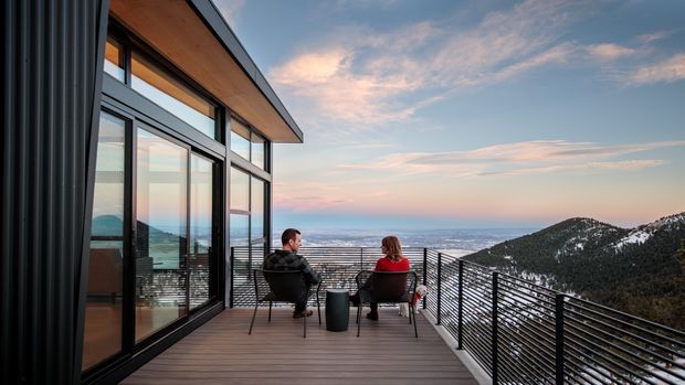 Modern mountain-view residence with extensive floor-to-ceiling glazing opening onto a timber deck. Two people sit on outdoor chairs overlooking a snow-dusted mountain ridge and expansive distant water or valley views. The building features dark vertical exterior cladding, black-framed glazing, a projecting roof edge, and horizontal metal cable or rail balustrades. The image has a dramatic sunset atmosphere.