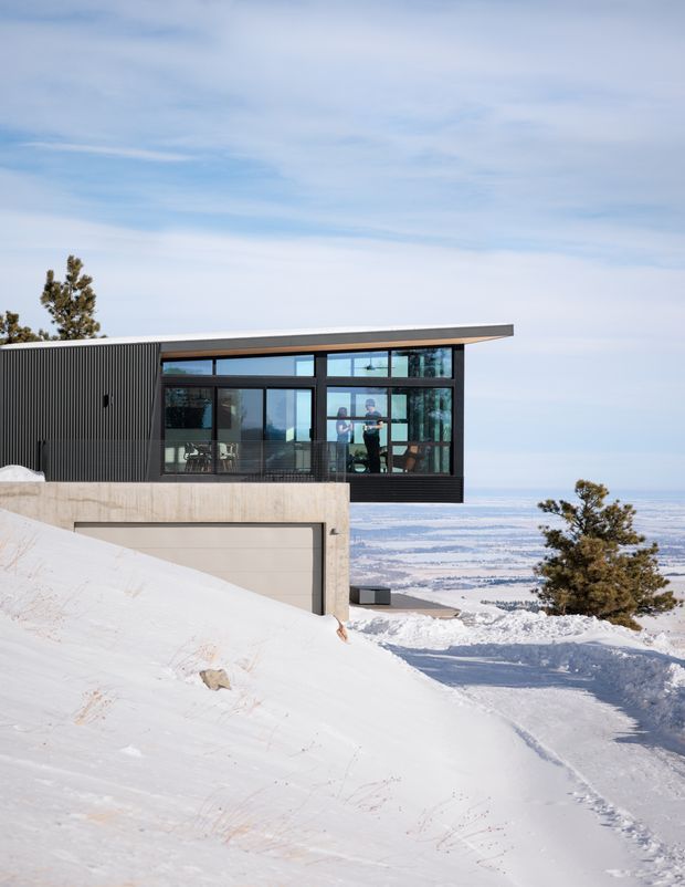 Exterior photograph of a contemporary flat-roofed house set on a steep, snow-covered alpine site. The building features dark vertical cladding, expansive black-framed glazing, a projecting upper volume, and a concrete garage structure below. Snowy mountains and a broad wintry landscape are visible beyond.
