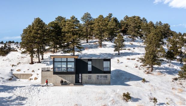 A compact modern cabin set into a snowy, forested hillside. The building has a dark vertical-clad upper volume, a concrete lower level, large floor-to-ceiling windows, and a low-pitched flat roof. Snow-covered terrain and pine trees surround the isolated alpine dwelling.