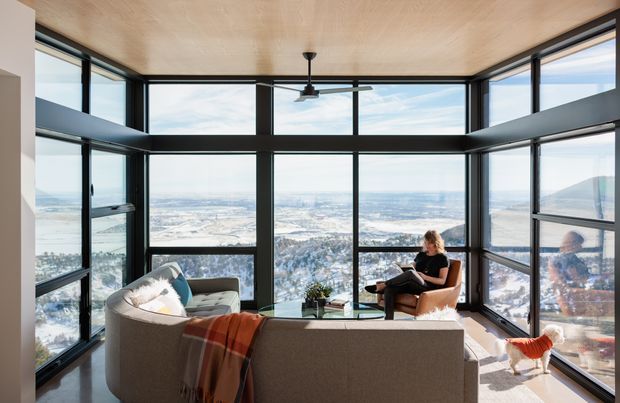 A bright contemporary living room positioned within expansive floor-to-ceiling corner glazing, offering panoramic views over a snowy landscape and distant water. The room features a large curved sectional sofa, a lounge chair, a small coffee table, a ceiling fan, and a warm timber ceiling. Black-framed windows create a strong structural grid around the space, while a person and a small dog are present.