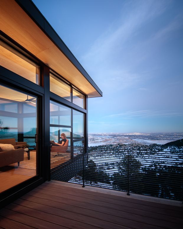 Modern multi-level home viewed from a timber deck terrace, featuring expansive black-framed glazing, warm timber soffits and interior finishes, and a horizontal metal balustrade. The elevated outlook overlooks a snow-covered mountainous or alpine landscape at dusk, with a living area visible through the open glazing.